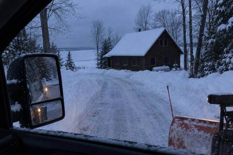 A freshly plowed driveway seen from a truck window at dusk, leading to a dark cedar cottage with no lights on and deep snow on its roof, a propane tank beside it and a frozen bay behind