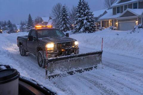 A pickup with a straight plow blade on a snow-covered subdivision street before sunrise, plowed banks along both shoulders and porch lights on in the houses behind
