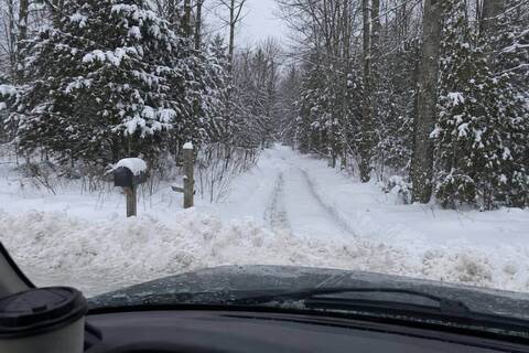 An unplowed two-track road under deep snow between bare hardwoods, a single set of tire ruts fading into the trees