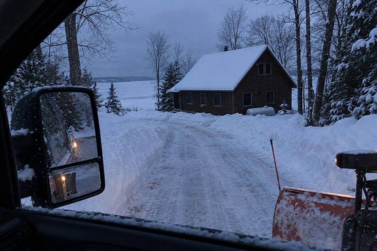 A freshly plowed driveway seen from a truck window at dusk, leading to a dark cedar cottage with no lights on and deep snow on its roof, a propane tank beside it and a frozen bay behind
